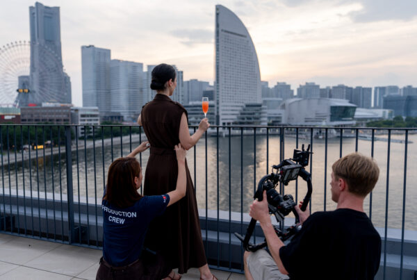 Woman in a brown dress holding an orange drink on a rooftop balcony as a film crew records her against a city skyline including a river and modern buildings.