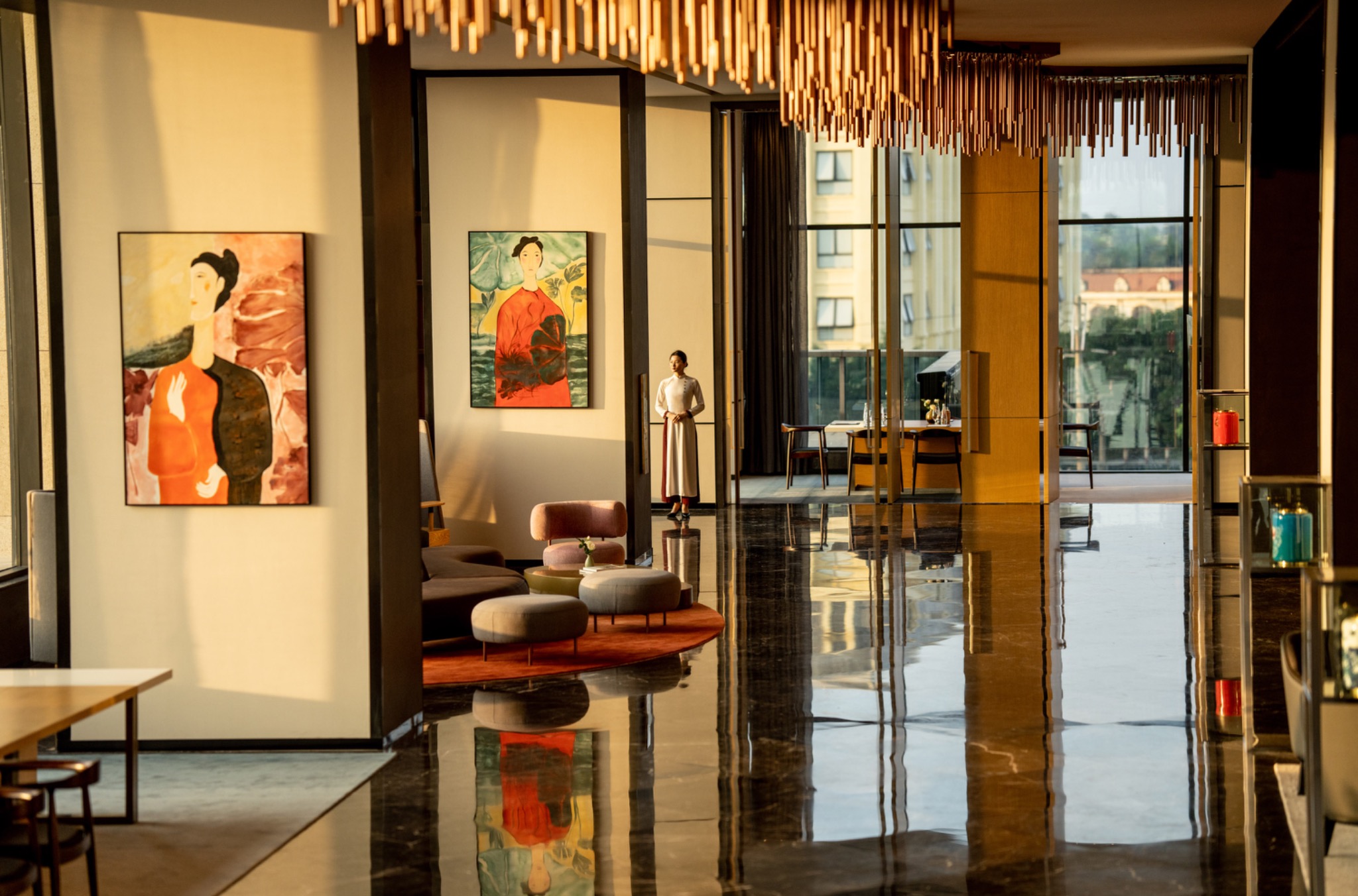 A woman stands in a sunlit hotel lobby with artwork, hanging lights and reflections across a polished floor.