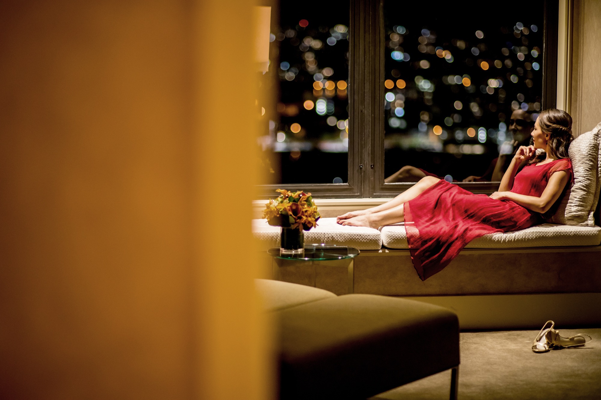 A woman in a red dress relaxes on a hotel window seat overlooking city lights at night.
