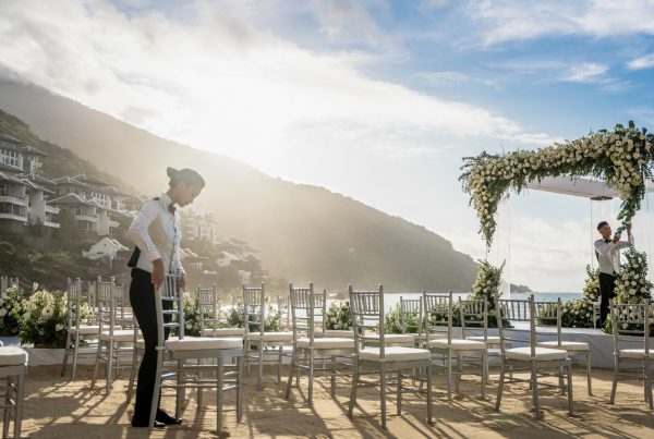 Hotel staff arrange chairs and a flower-covered arch for a beach wedding beside a hillside resort.