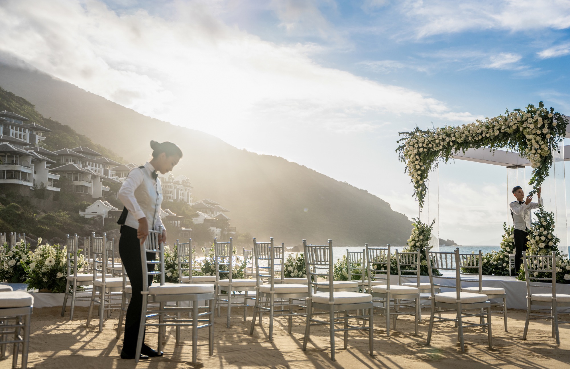 Hotel staff arrange chairs and a flower-covered arch for a beach wedding beside a hillside resort.
