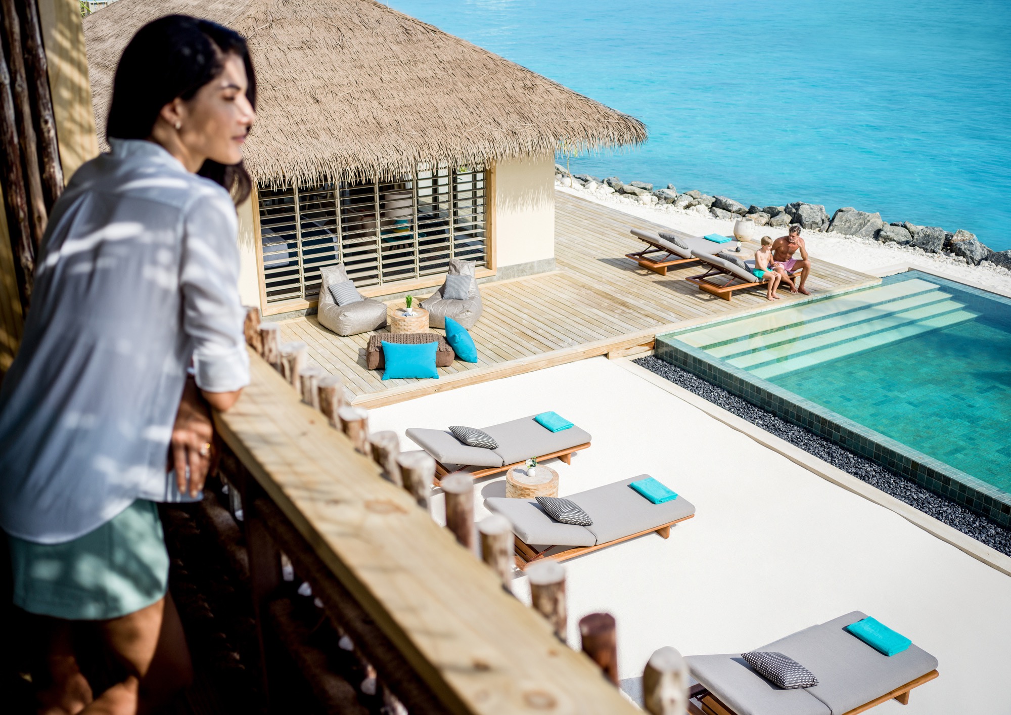 A woman looks from a balcony toward a man and child beside a private pool overlooking turquoise sea.