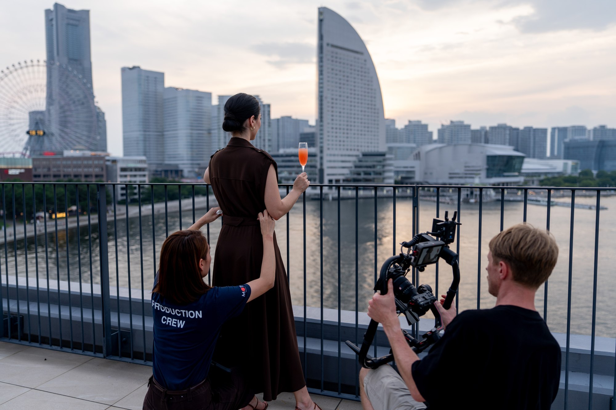 A camera operator and crew member film a model holding a drink on the hotel rooftop with Yokohama behind her.