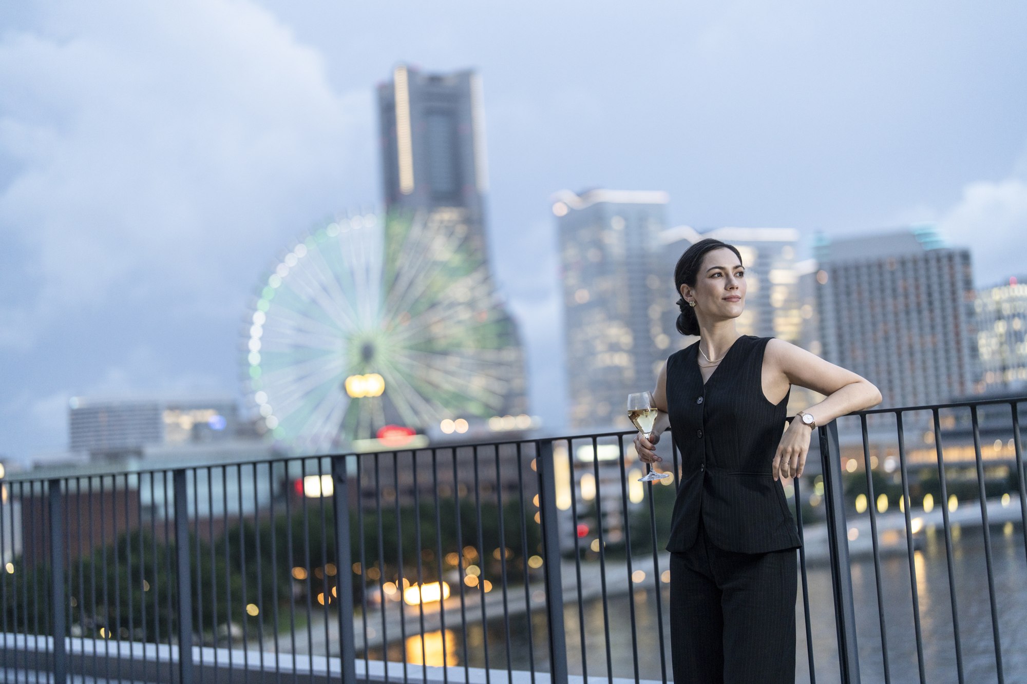 A woman stands on the hotel rooftop with the Yokohama skyline behind her at dusk.